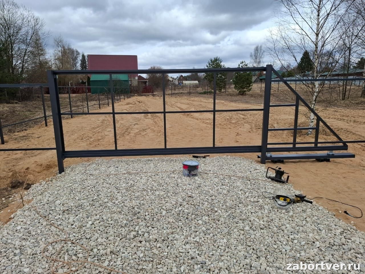 Черный металлический каркас откатных ворот на стадии монтажа, установленный на гравийной подушке с инструментами на переднем плане