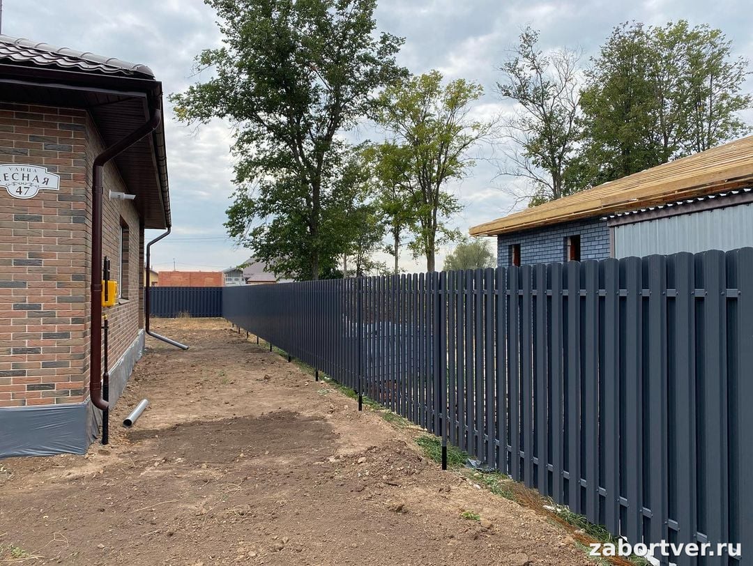 Сплошной забор из темного металлического евроштакетника цвета RAL 7024, установленный между кирпичными домами