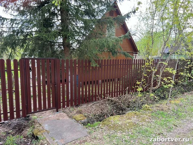 Глухой забор из вертикального металлического штакетника темно-коричневого цвета с распашной калиткой на фоне деревянного дома