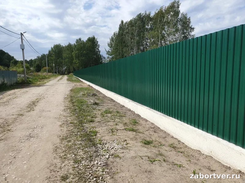 Длинный сплошной забор из зеленого профнастила на белом бетонном фундаменте вдоль грунтовой дороги