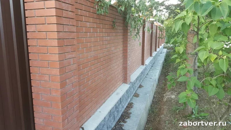 Готовый кирпичный забор из красного облицовочного кирпича с выделенными столбами и цоколем из серого камня