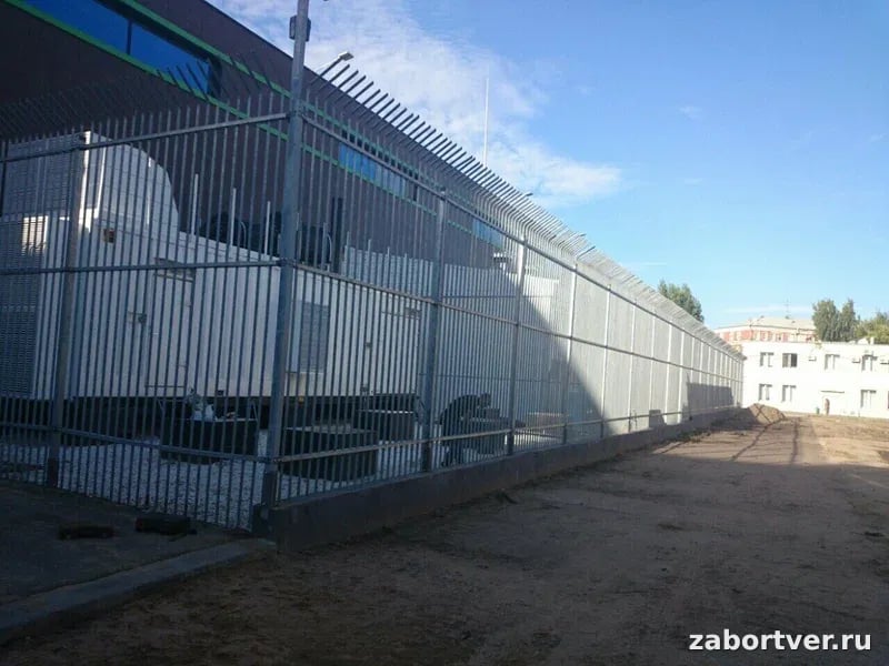 Высокий оцинкованный сварной забор с защитным наклонным козырьком и шипами, ограждающий промышленное оборудование