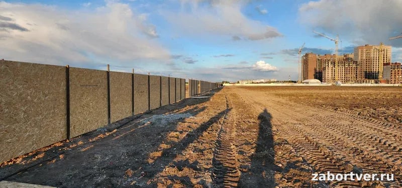 Длинный временный забор из плит OSB на металлических столбах, ограждающий строительную площадку с башенными кранами на фоне