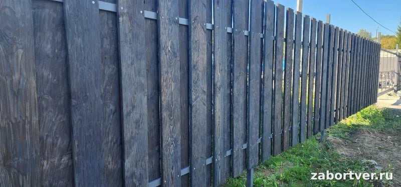 Глухой деревянный забор, окрашенный в темно-серый цвет, с вертикальными досками, закрепленными на металлических лагах.