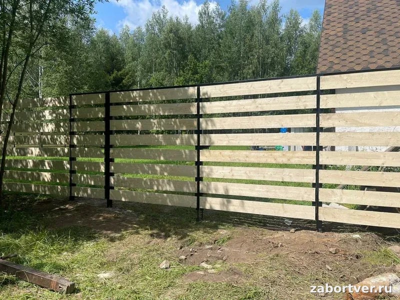 Современный горизонтальный забор из светлого дерева на черном металлическом каркасе, установленный на участке с зеленым газоном.