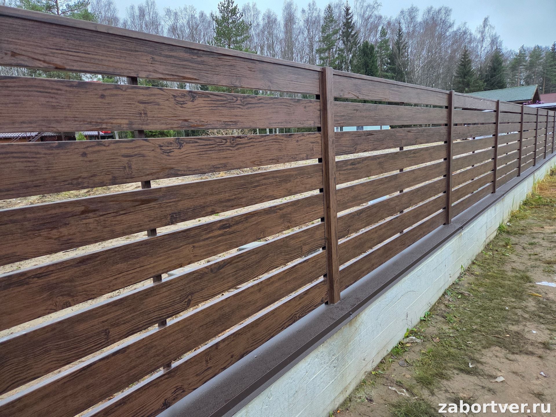 Горизонтальный металлический забор с ламелями под темное дерево на белом бетонном цоколе