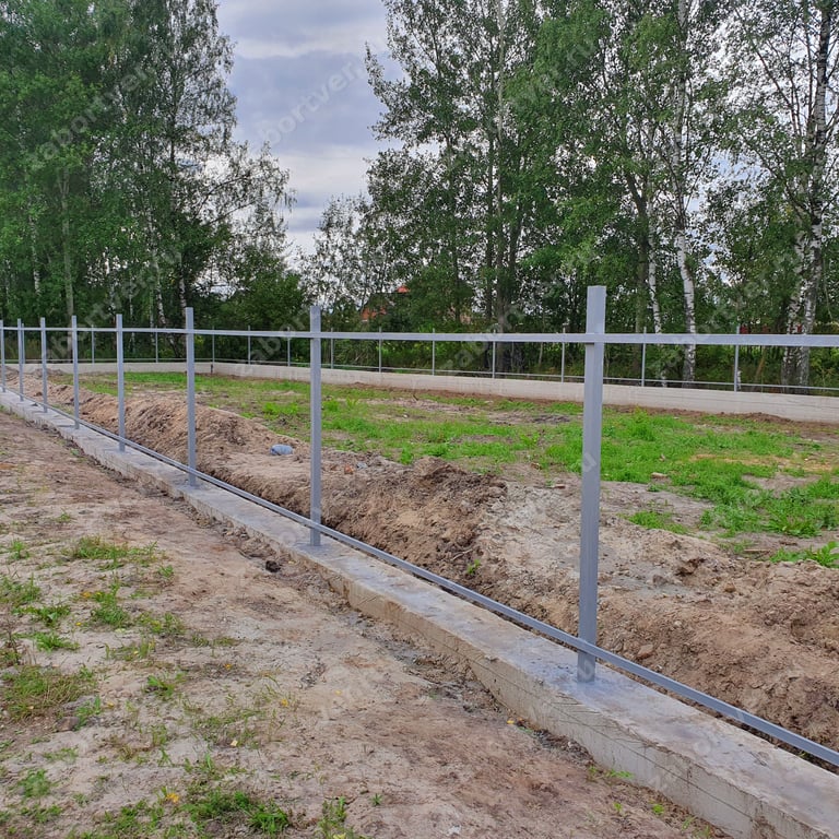 На фотографии запечатлен процесс монтажа металлического каркаса забора из профильной трубы на монолитном ленточном фундаменте. Видны установленные вертикальные столбы и приваренные горизонтальные лаги серого цвета на фоне земельного участка с деревьями.