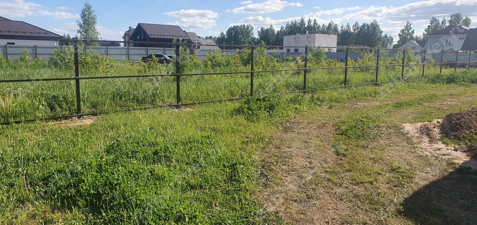 На фотографии представлен металлический забор типа 'ранчо', выполненный из профильной трубы с горизонтальным расположением перекладин. Ограждение темного цвета установлено на границе зеленого участка, на заднем плане видны соседние дома и другие заборы.