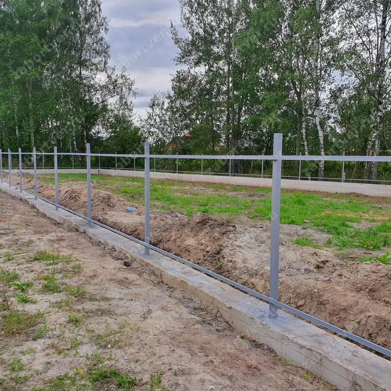 На фотографии запечатлен процесс монтажа металлического каркаса забора из профильной трубы на монолитном ленточном фундаменте. Видны установленные вертикальные столбы и приваренные горизонтальные лаги серого цвета на фоне земельного участка с деревьями.