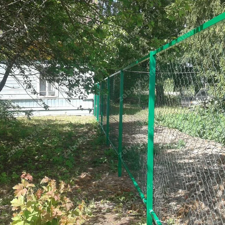 Классический забор из сетки-рабицы, установленный на металлические столбы зеленого цвета. Конструкция дополнена натяжной проволокой для предотвращения провисания сетки.