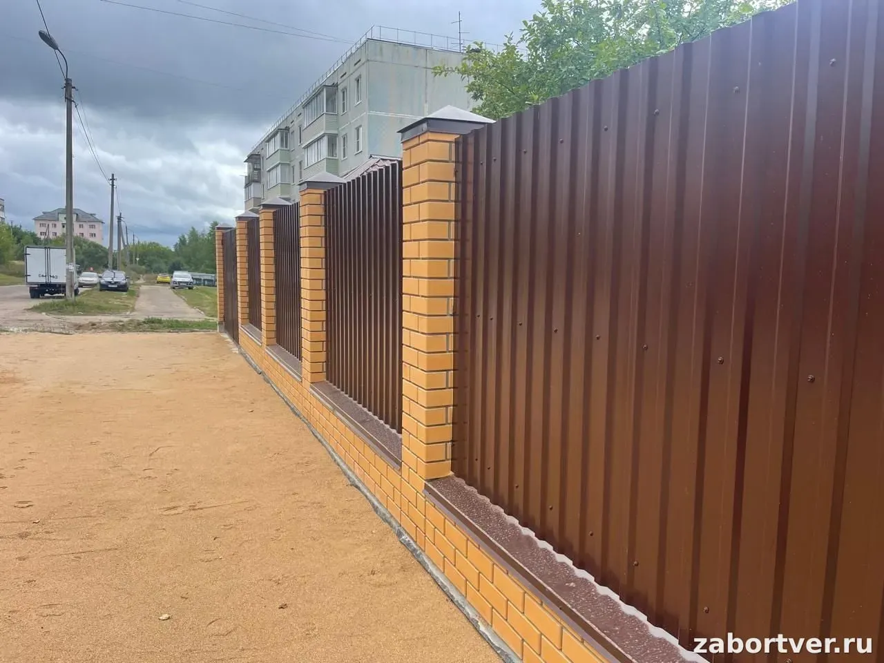 Это сплошной забор, секции которого выполнены из коричневого профнастила, установленный на кирпичном основании с кирпичными столбами светло-желтого цвета. Стиль ограждения — современный и прочный, обеспечивающий хорошую защиту от посторонних глаз.