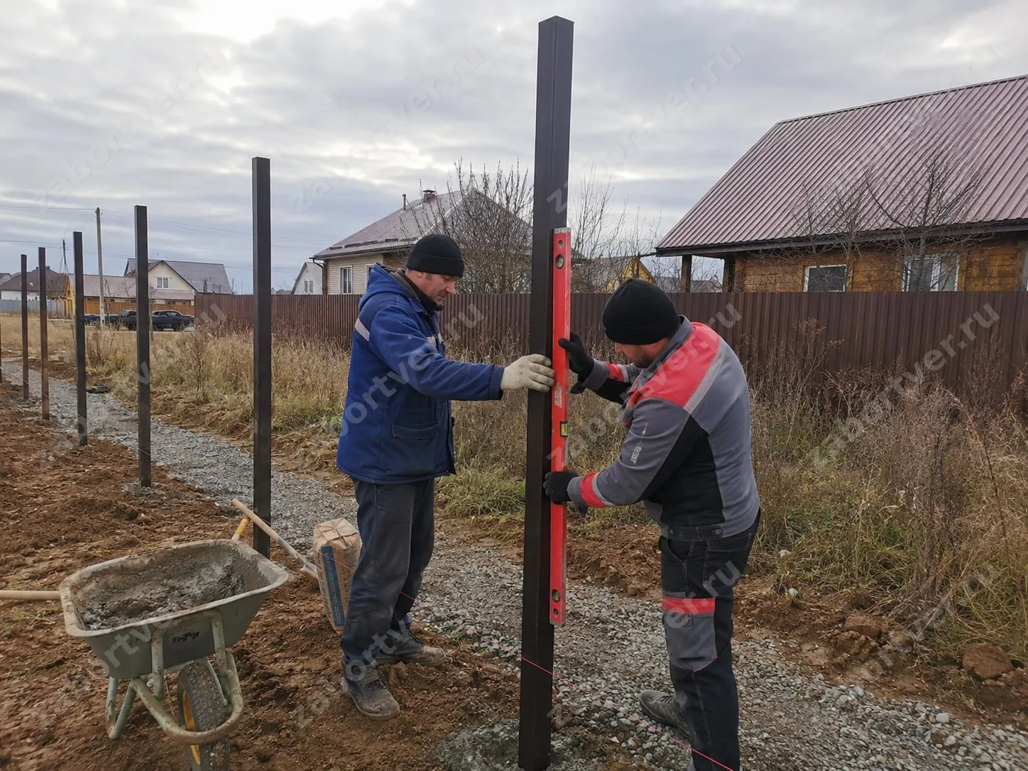 На фотографии запечатлен процесс установки забора: двое рабочих выравнивают по уровню и бетонируют металлические столбы из профильной трубы. На заднем плане виден уже установленный забор из профнастила, что указывает на тип возводимого ограждения.